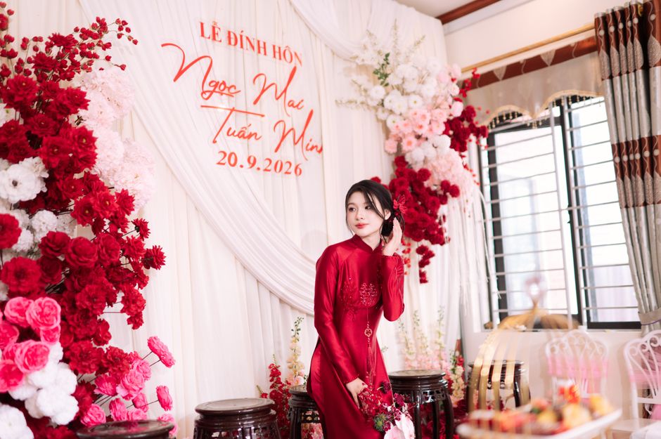 A young woman in a traditional red Vietnamese áo dài dress is standing indoors amidst a floral arrangement backdrop featuring pink, white, and red flowers, including roses and delphiniums, arranged 