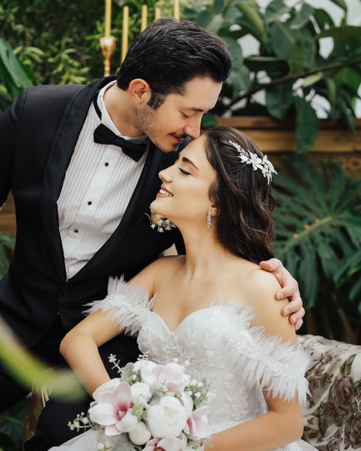 A bride and groom share a joyful moment indoors amidst lush green tropical plants. The bride, wearing a white wedding gown with feathered off-shoulder details and a sparkling tiara, holds a floral bou