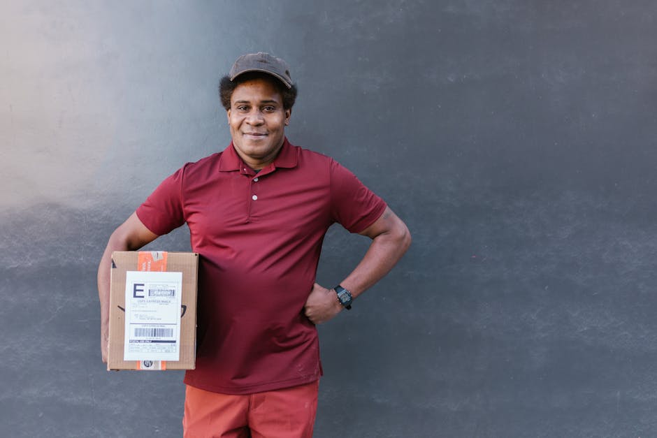 A smiling delivery person of African descent wearing a maroon polo shirt, matching maroon apron, and a dark cap, holding a small rectangular cardboard parcel with a shipping label in front of a plain 