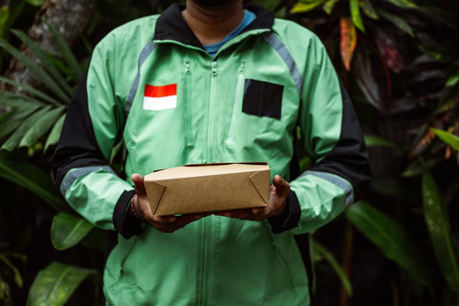 A person wearing a bright green and black jacket with a red and white flag patch on the chest, holding a rectangular brown cardboard box with folded edges. The individual is standing outdoors surround