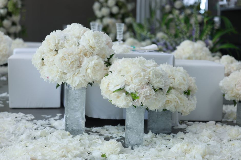 Two floral arrangements featuring large, lush white carnations and roses in tall, clear glass vases placed on a surface sprinkled with white petals. The arrangements are densely packed with fresh, tex
