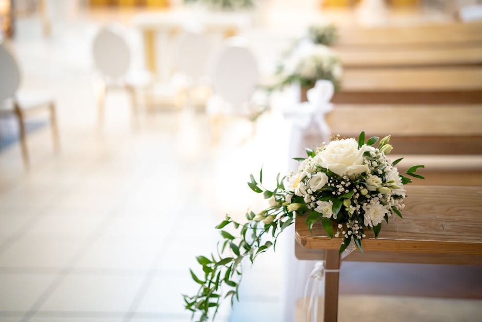 A delicate wedding flower arrangement featuring white roses, lisianthus, and baby's breath with lush green foliage, placed on a wooden church pew. The bouquet is freshly arranged with vibrant petals a