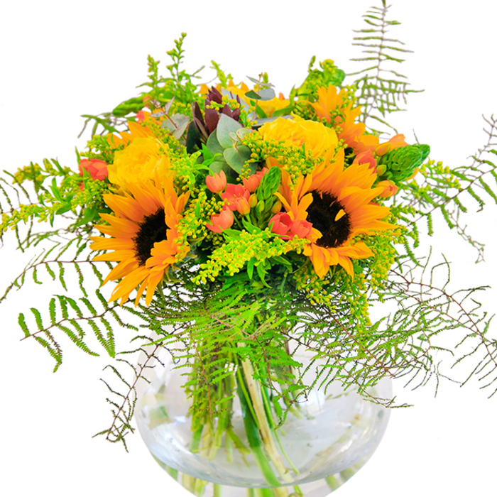 The first impression this bouquet leaves is one of pure, joyous energy inspired by the heart of Wimbledon. Golden petals of sunflower and garden rose surge outward from a crystal-clear, round glass vase, their yellow hues as vibrant as sunlight dancing across Wimbledon Village Green. Interlaced among the bold blooms, clusters of cherry-red hypericum berries glisten like tiny treasures, creating a delightful contrast to the soft matte of the petals. Cascades of lush fern fronds and light, airy sprigs-possibly goldenrod-spill outward, imbuing the arrangement with texture and a sense of natural movement reminiscent of breezy afternoons by the River Wandle. Silvery eucalyptus foliage peeks through, grounding the bouquet with a subtle, aromatic freshness. Viewed up close, you'd feel the intricate interplay of textures: the velvety softness of the sunflower faces, the densely layered, unfolding roses, the crisp snap of youthful greenery. The water-filled vase not only displays the interwoven stems but magnifies their verdancy, enhancing the arrangement's lively character. This bouquet's essence echoes celebrations held in local halls and cafes, catching the golden light and festive spirit that defines many a Wimbledon gathering. Whether it graces a birthday table, marks a heartfelt anniversary, or offers a spontaneous burst of happiness in someone's home, it radiates warmth and a sense of open-hearted celebration-making it a perennial favourite among our community's floral gifts.